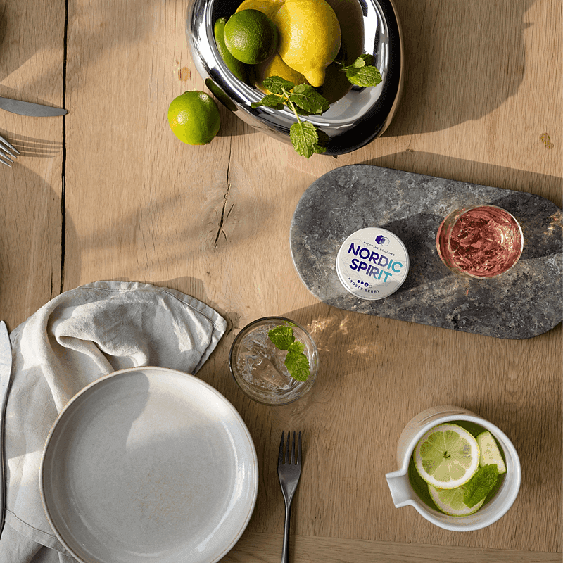 Top down shot showing a can of Nordic Spirit Frosty Berry Nicotine pouches on a table with fresh sliced fruit and cocktail drinks.