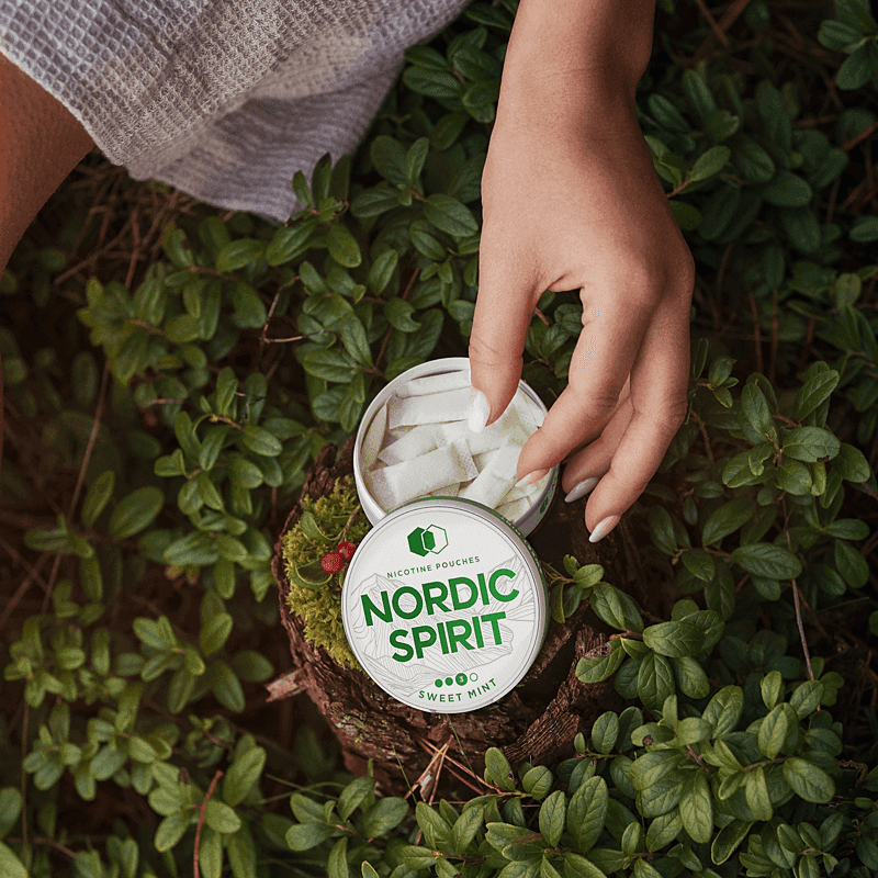 Lifestyle shot showing an open can of Nordic Spirit Sweet Mint Nicotine pouches with a female hand reaching in to pick up a pouch.